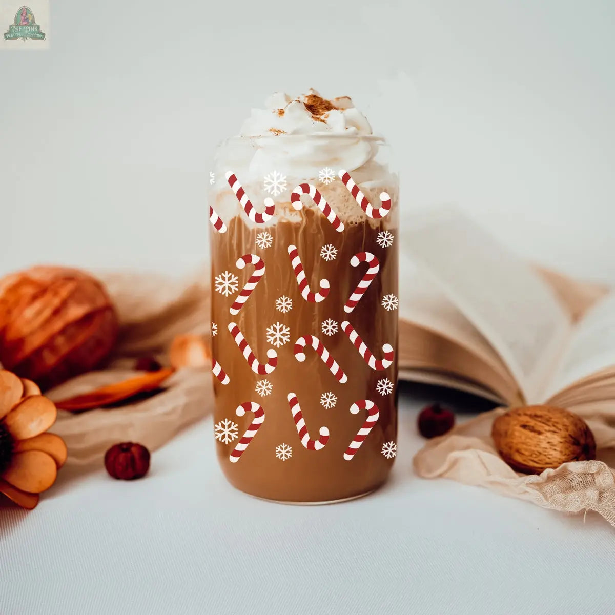 A festive Christmas cup of coffee topped with whipped cream and cinnamon sits on a white surface decorated with a Candy Cane and snowflake patterns, alongside an open book, a pumpkin, a walnut, and autumn flowers.