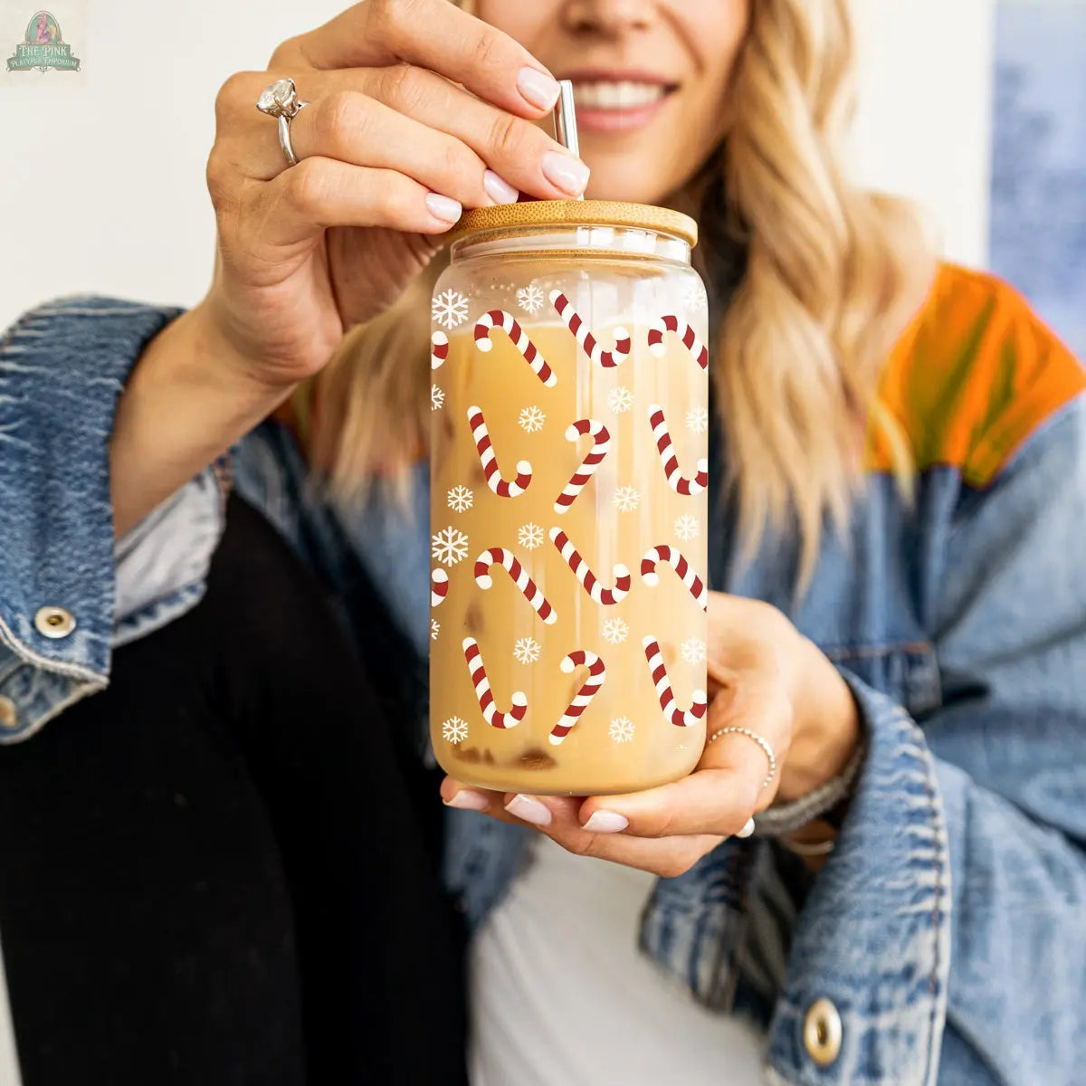 A woman in a denim jacket and engagement ring holds the Candy Cane holiday glass cup, decorated with candy canes and snowflakes, filled with a creamy iced drink and topped with a metal straw.