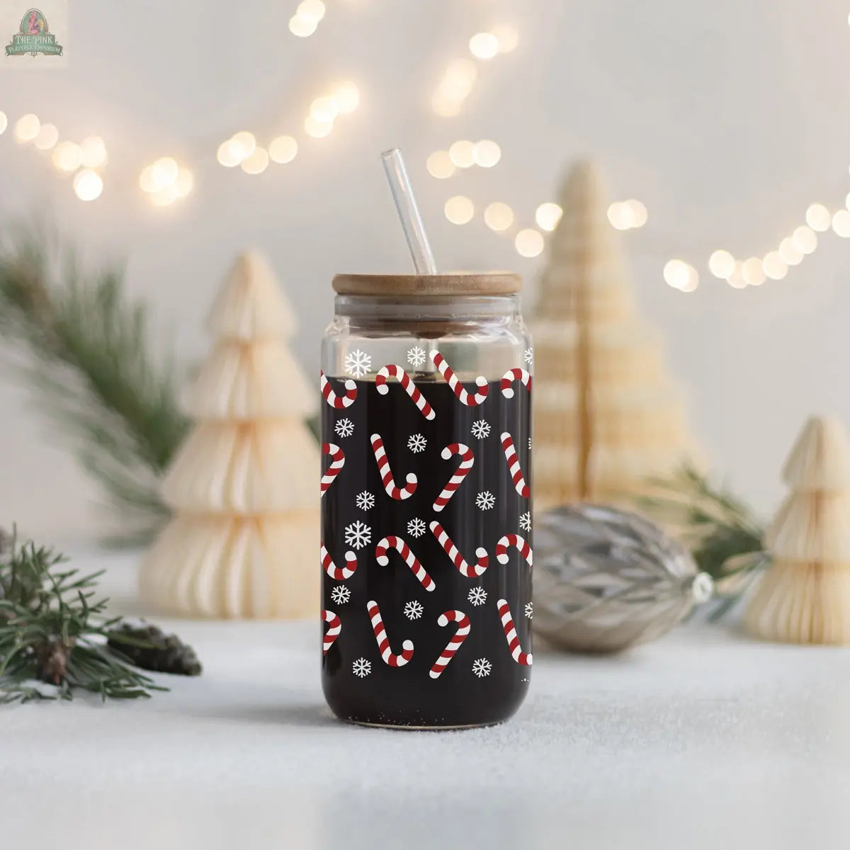 The Candy Cane cup features a wooden lid and straw, with candy cane and snowflake designs. It sits on a snowy surface, with blurred holiday lights and Christmas trees glowing warmly in the background.