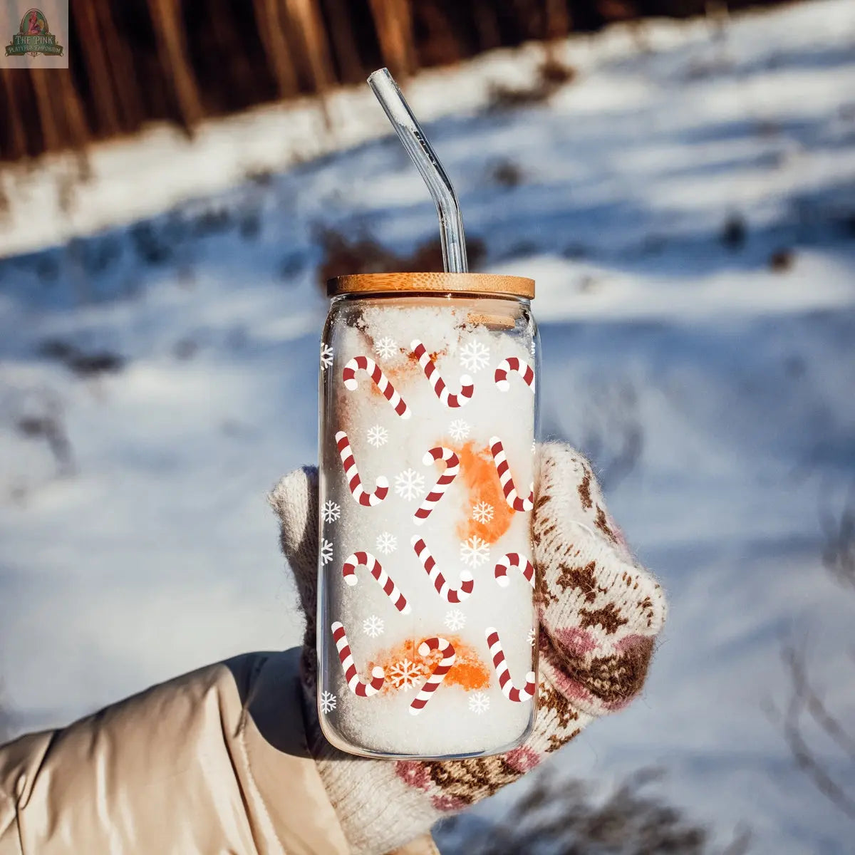 Wearing patterned winter gloves, a person holds the Candy Cane Christmas cup with a bamboo lid and glass straw—decorated with candy canes and snowflakes—in a snowy outdoor setting.