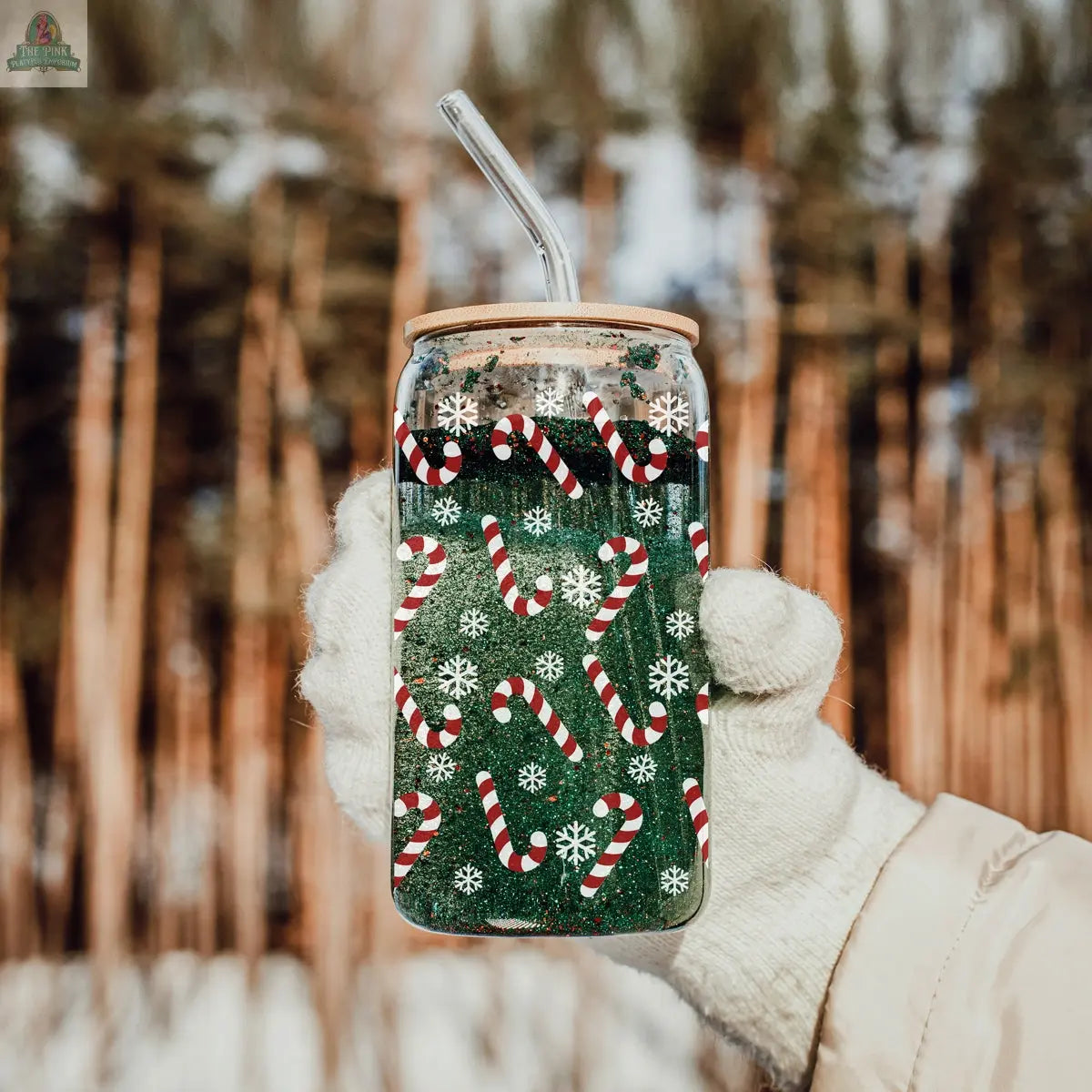 A gloved hand holds a Candy Cane Christmas cup with a clear straw, decorated with snowflakes and candy canes and filled with a green drink, while snow-covered trees blur in the background.