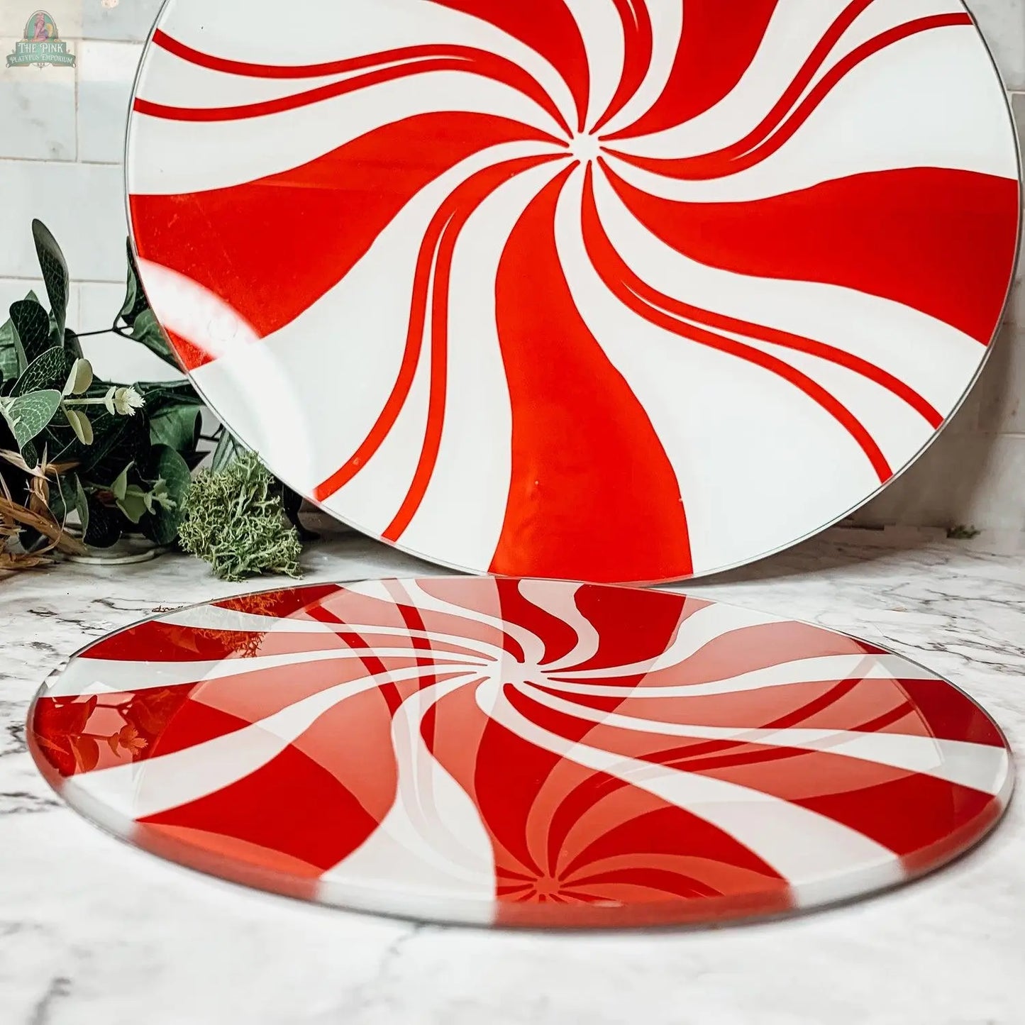 Displayed on a marble countertop with green leafy accents, the Candy Cane Serving set features two round glass platters with red and white peppermint swirl patterns—perfect for festive holiday kitchen decor.