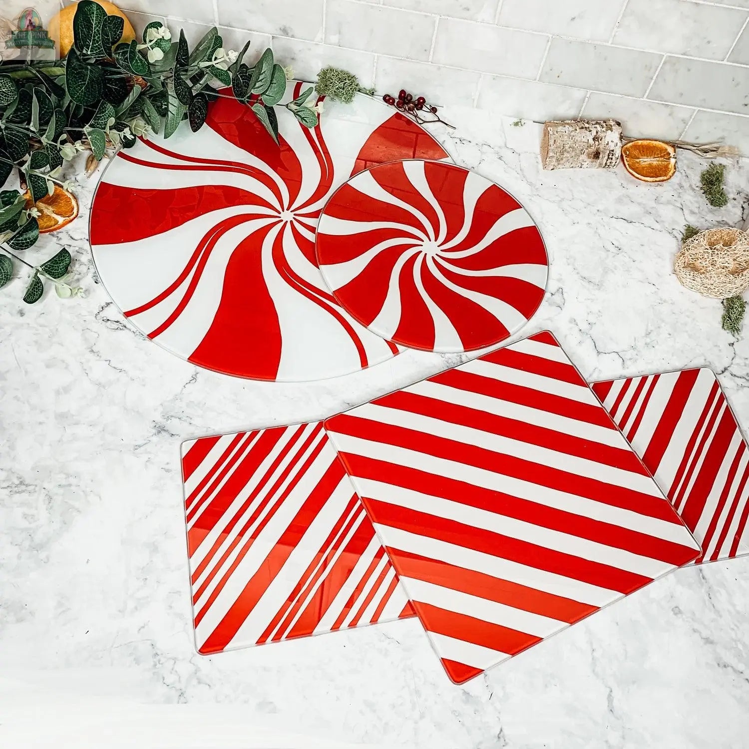 The Candy Cane Serving set features two round plates with red and white peppermint swirls and three square plates with diagonal stripes, creating a festive holiday display on a marble countertop with greenery and dried orange slices.
