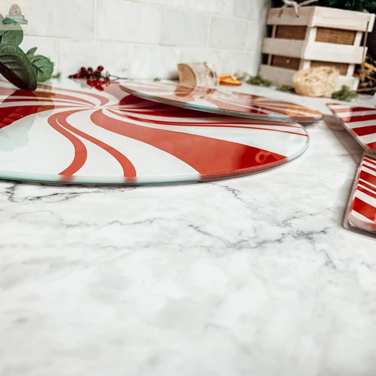 A close-up of Candy Cane Serving platters featuring red and white peppermint swirl designs rests on a marble countertop, accented with greenery and festive holiday kitchen decor in the background.