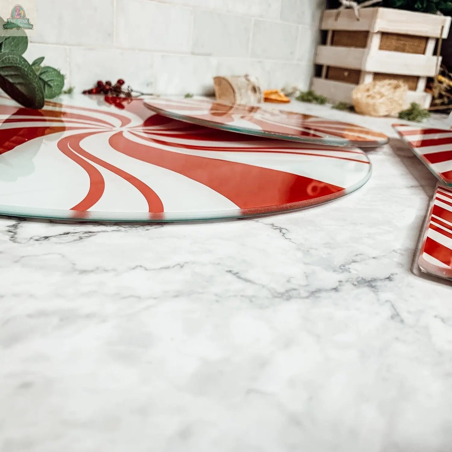 A close-up of Candy Cane Serving platters featuring red and white peppermint swirl designs rests on a marble countertop, accented with greenery and festive holiday kitchen decor in the background.