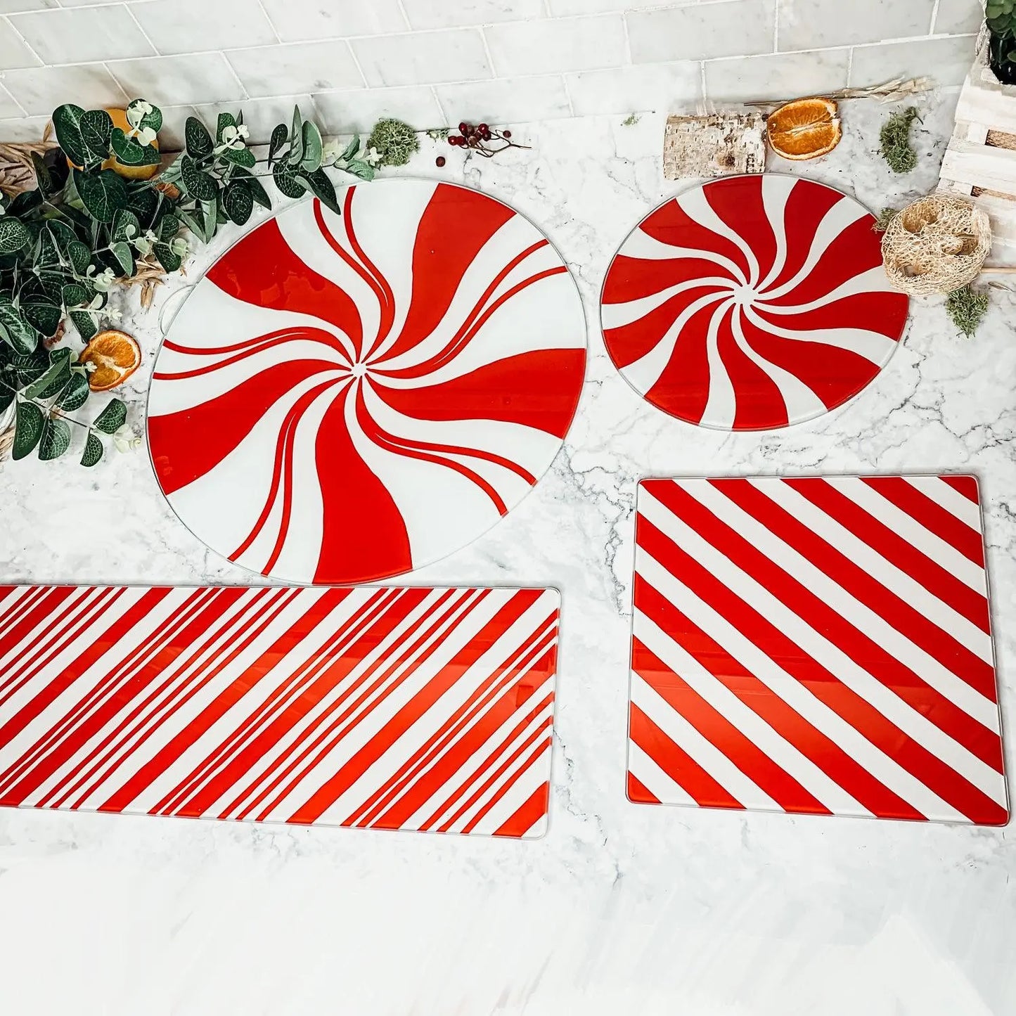 The Candy Cane Serving set features four glass platters with red and white peppermint swirl and stripe designs, displayed on marble amid greenery, dried oranges, and rustic holiday kitchen accents.