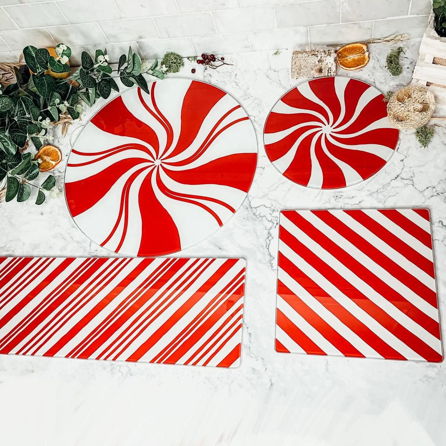 The Candy Cane Serving set features four glass platters with red and white peppermint swirl and stripe designs, displayed on marble amid greenery, dried oranges, and rustic holiday kitchen accents.