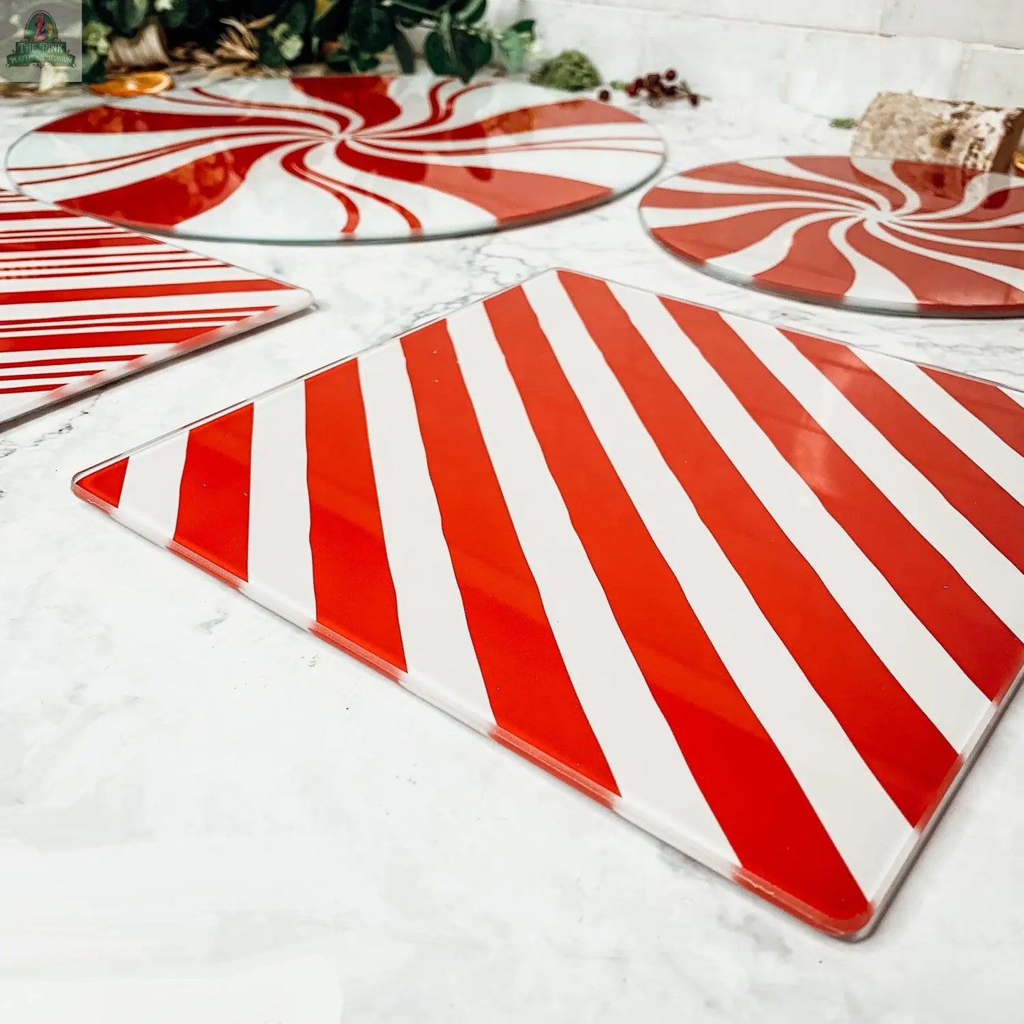 Candy Cane Serving glass plates and platters, hand painted with red and white peppermint stripes, are displayed on marble, bringing festive flair to your kitchen decor with greenery in the background.