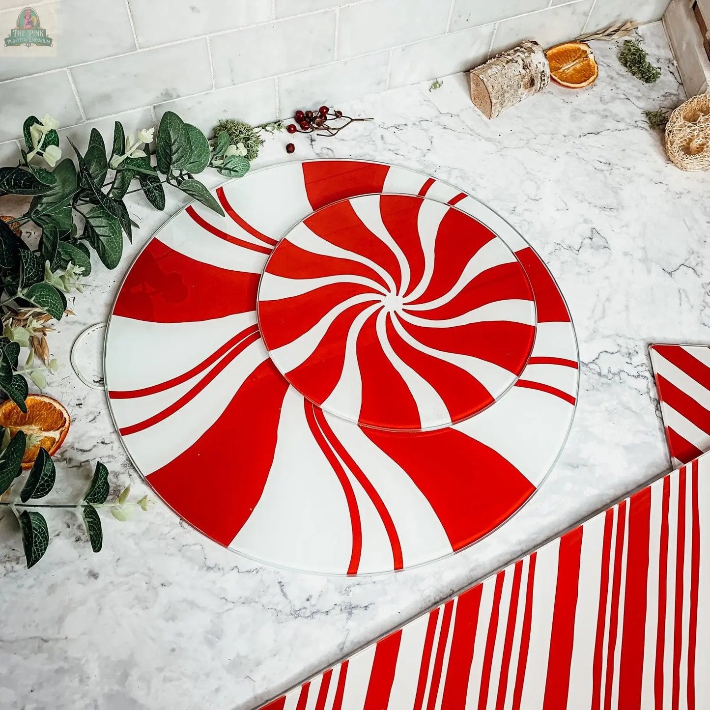 The Candy Cane Serving, a round, spiral-patterned red and white tray from our hand painted glass collection, is displayed on marble with greenery, dried orange slices, and festive holiday kitchen decor.