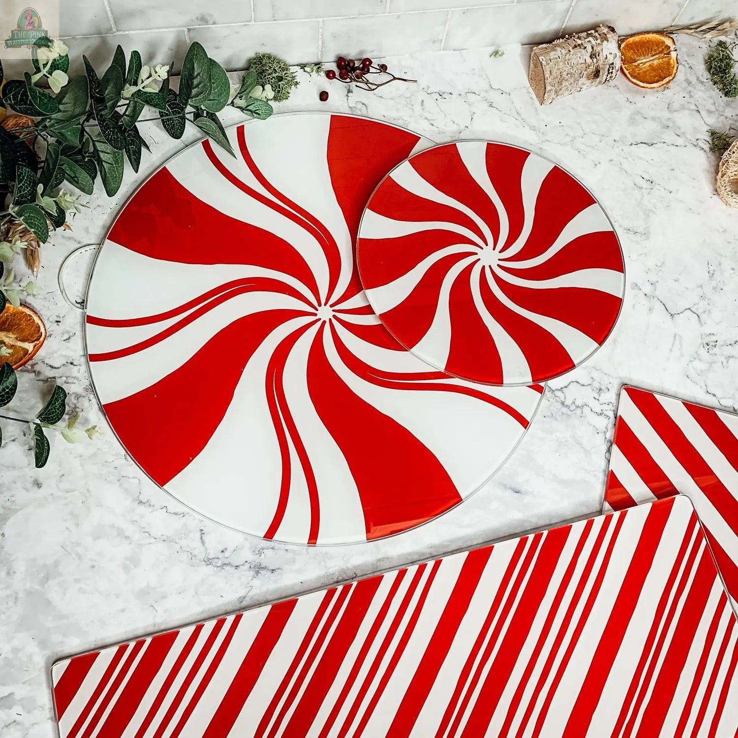The Candy Cane Serving collection features two round plates with red and white peppermint swirls and a rectangular platter with matching stripes, hand painted for festive holiday kitchen decor. Displayed with marble, greenery, and dried orange slices.