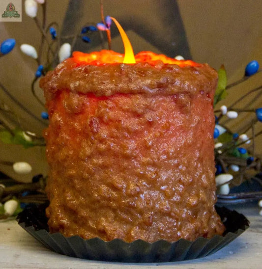 The Caramel Coffee Cake Electric Candle, with a textured reddish-brown design and warm glow, sits in a black fluted holder. Decorative stems featuring blue, white, and brown buds are visible in the background.