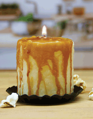 The Caramel Corn Mini Hearth candle features caramel sauce-like wax drips on a black holder, accented with popcorn pieces on a wooden base—a charming rustic decor piece for your cozy space.