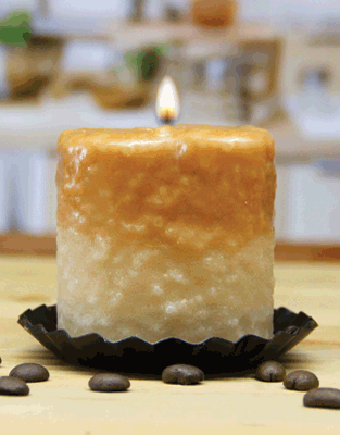 The Caramel Latte Mini Hearth candle by Pink Platypus Emporium features brown and white paraffin wax, lit on a black tray with coffee beans scattered on a light wooden surface. Blurred kitchen background. Made in the USA.