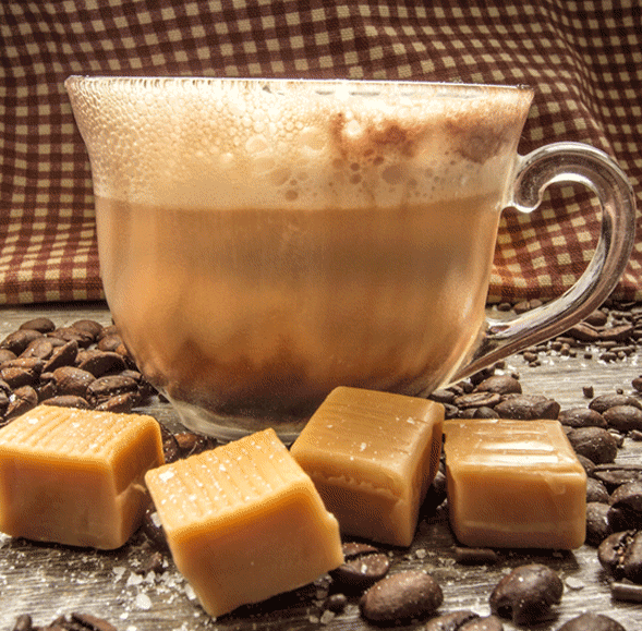 A clear glass cup of frothy coffee sits on a wooden surface with coffee beans, four caramel candies, and the Caramel Latte Mini Hearth candle by Pink Platypus Emporium. A brown checkered cloth is in the background.