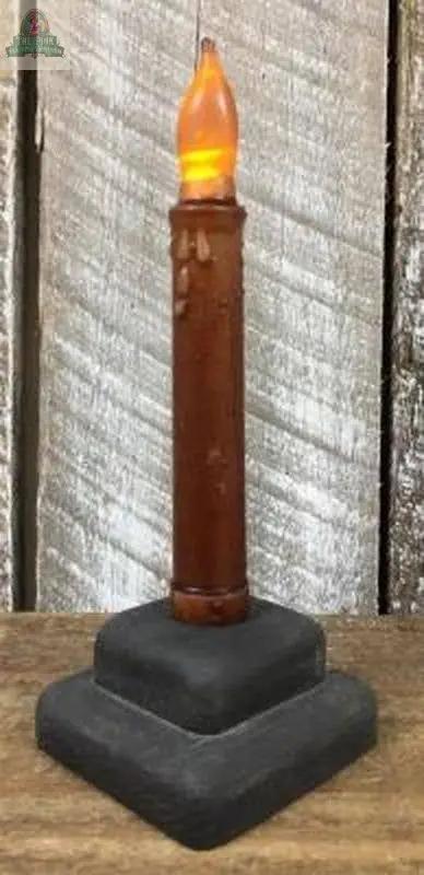 The Caramel Taper LED Candle w/Timer features a glowing orange flame, rustic design, two-tiered black base, and is set against a weathered wooden background.