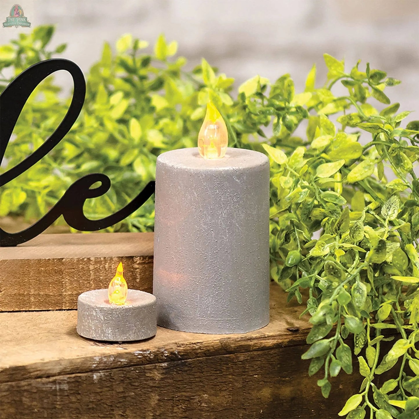 Two flameless LED candles, including a Cement Look Timer Pillar 2.25 x 4, sit on wood among green plants. Each battery-operated candle features a realistic cement design and charming black script, perfect for farmhouse decor.