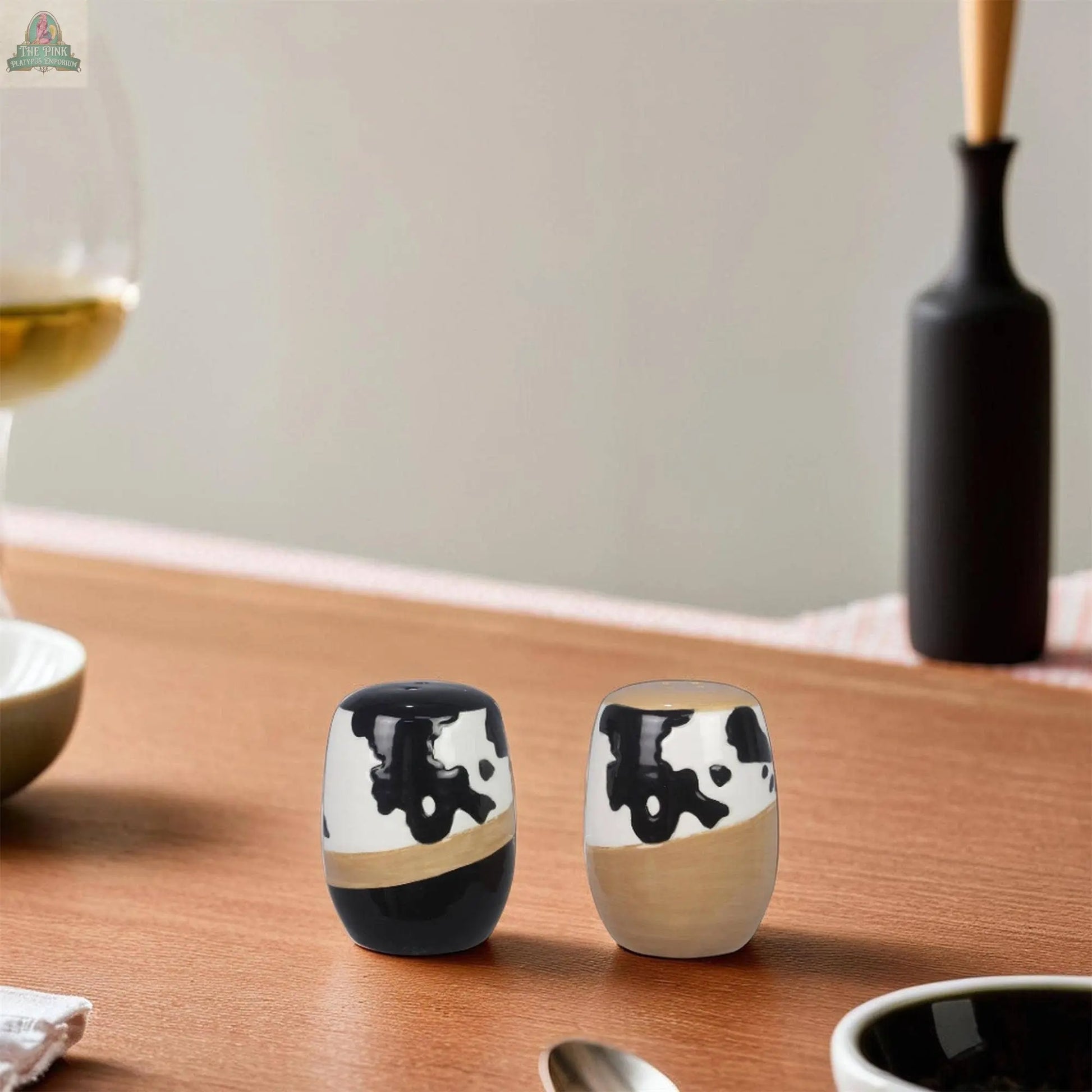 Young's Ceramic Country Salt Pepper Shaker Set Cow Print features two small shakers with black and white cow print designs, displayed on a wooden table amid dishes, with a wine glass and black vase in the blurred background.