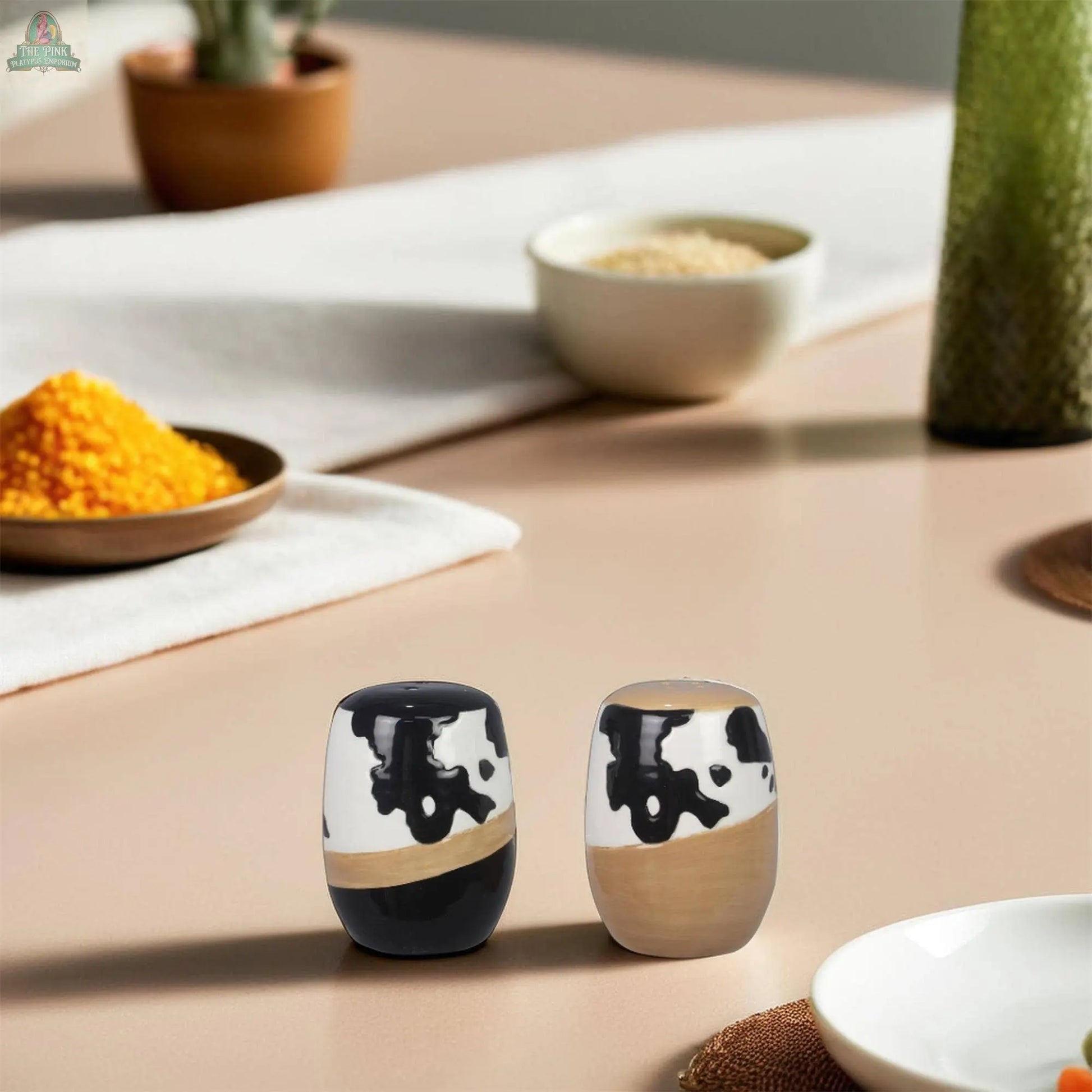 The Young's Ceramic Country Salt Pepper Shaker Set features a modern cow print design in black and beige, sitting on a light table. These stylish shakers complement farmhouse kitchen decor, with bowls of spices and a white runner behind them.