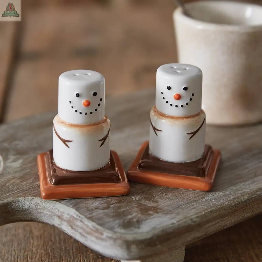 The Ceramic Smores Salt and Pepper Shakers, shaped like smiling snowmen with orange carrot noses on brown bases, add a playful touch to your dining experience when displayed on a wooden board.