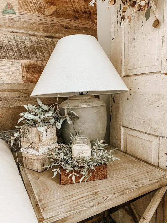 A rustic handmade wood nightstand holds a gray ceramic lamp, a small potted plant, a Cheese Mold - 1 Hole, Candle Pour Ready, Handmade #A - 6.5 with candle in glass jar, plus a decorative book with greenery and twine against a wood-paneled wall.
