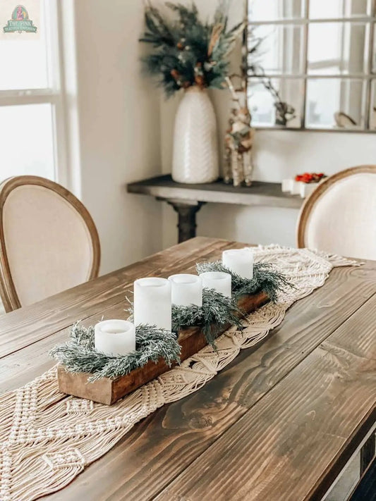 A rustic wooden dining table features a woven runner, four white candles with greenery, and a Cheese Mold - 5 Hole, Farmhouse Decor #B - 6.5 as decor. Soft beige chairs surround the table; pine branches in a vase adorn the console behind.