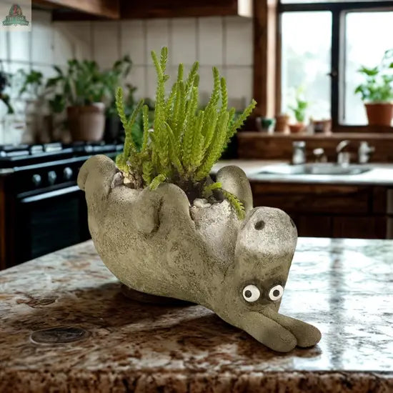 Chester, the Donkey Blobhouse Planter Laying by Pink Platypus Emporium—an upside-down donkey with googly eyes—holds a small plant on a kitchen counter, adding whimsical garden decor among potted plants and a sunny window.