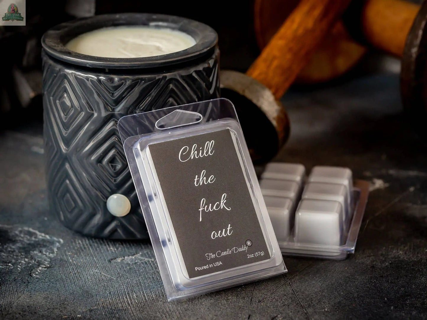 A black candle warmer and a pack of Chill the Fuck Out - Eucalyptus Mint Scented Melt rest on a dark surface, with dumbbells in the background.