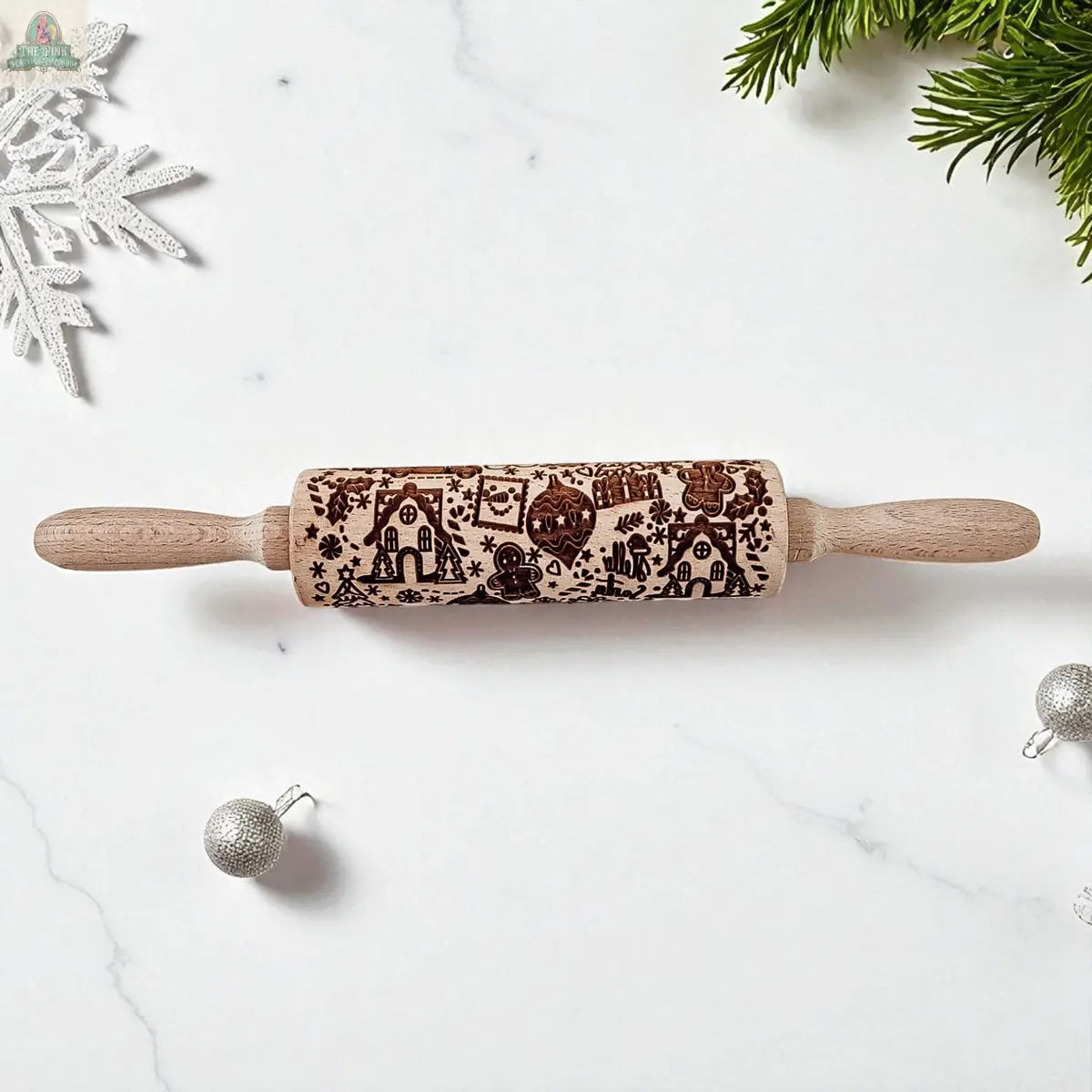The Christmas Rolling Pin Set | 10 Styles, featuring gingerbread houses, trees, and snowflakes, sits on white marble with festive silver ornaments and pine branches—an ideal holiday gift for bakers.