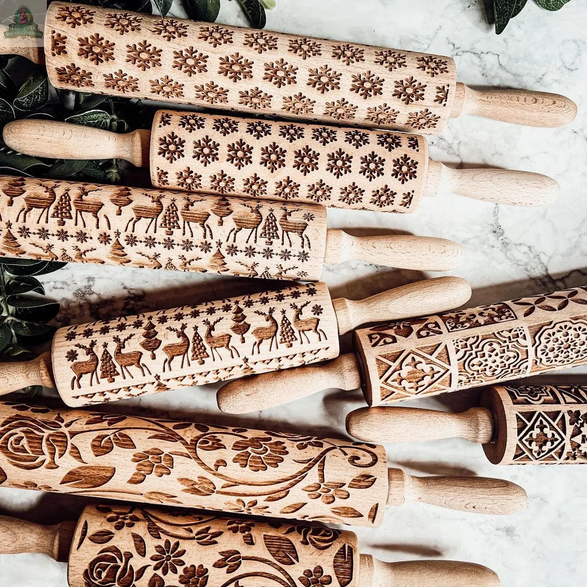 The Christmas Rolling Pin Set | 10 Styles features intricately engraved wooden pins in floral, snowflake, geometric, and festive patterns, arranged on marble with green leaves—ideal for bakers or pairing with your favorite cookie cutters.