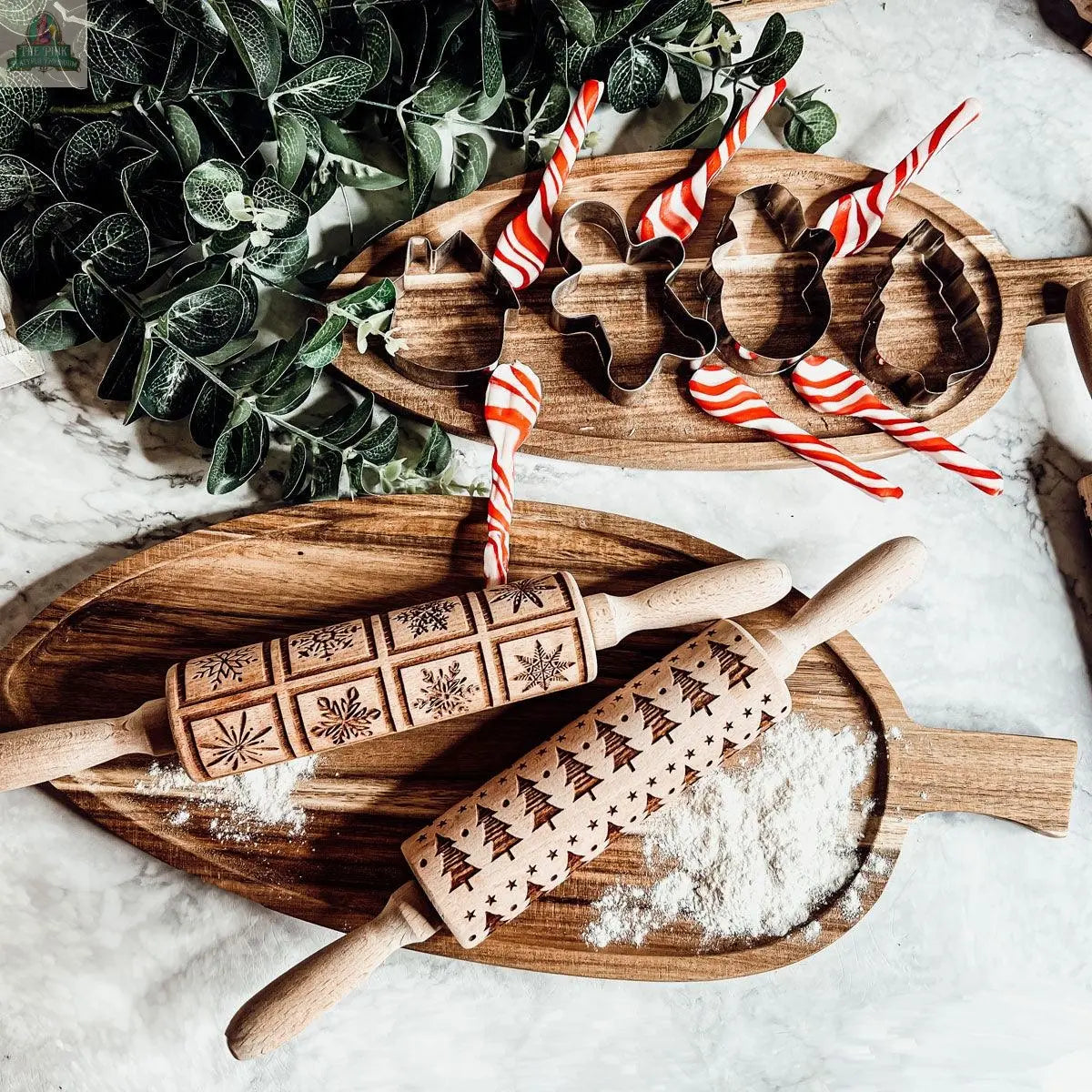 The Christmas Rolling Pin Set | 10 Styles rests in a wooden tray, dusted with flour, alongside festive cookie cutters and red and white striped stir sticks. Green leafy plants in the corner add cheer—making it a perfect gift for bakers.