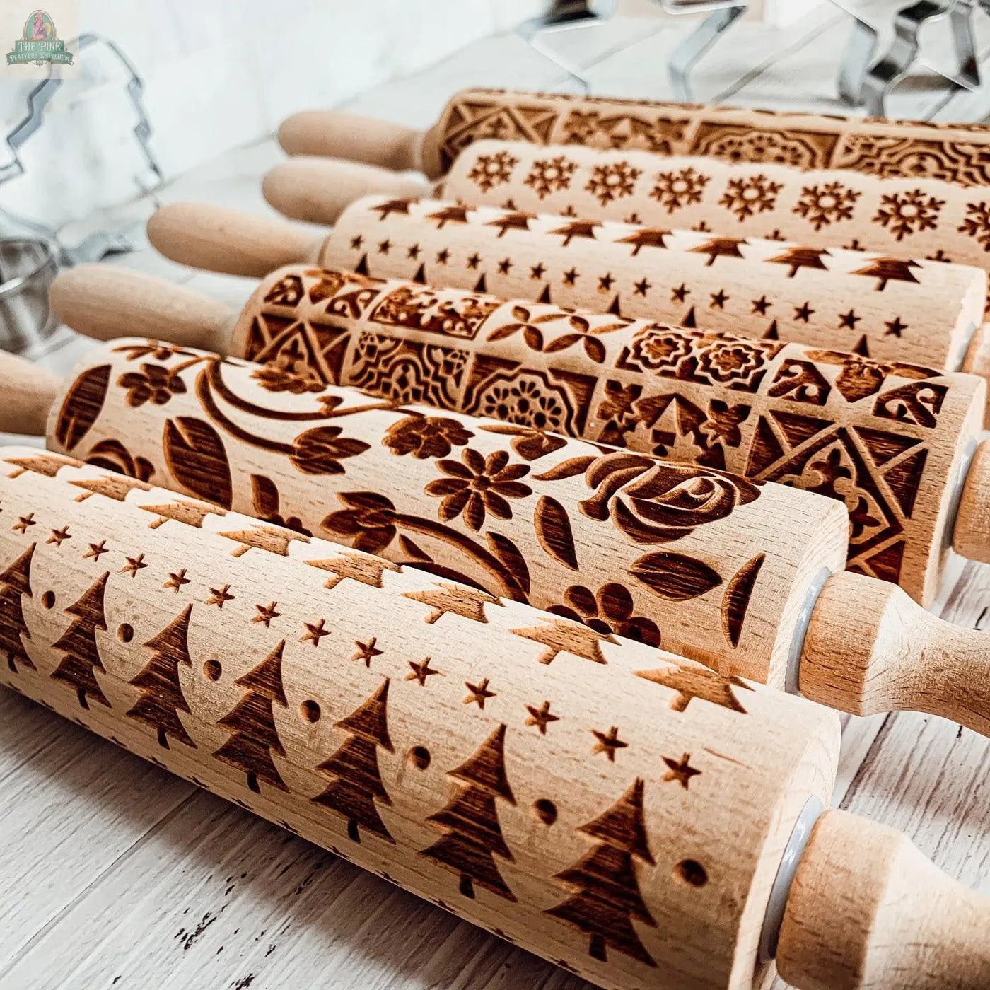 The Christmas Rolling Pin Set | 10 Styles features engraved holiday and floral patterns, shown lined up on a light wooden surface with cookie cutters in the background—an ideal gift for creative bakers.