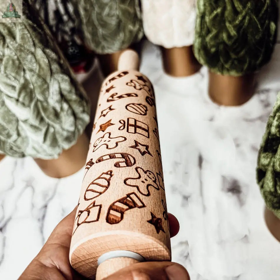 A hand holds a Christmas Rolling Pin from the Christmas Rolling Pin Set | 10 Styles, engraved with festive designs. In the background, textured green, white, and beige objects—possibly cookie cutters—sit on a marble surface.
