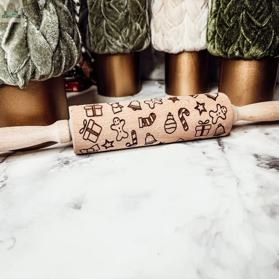 The Christmas Rolling Pin Set | 10 Styles features engraved festive designs and rests on a marble surface with green and tan velvet Christmas tree decorations in the background—a perfect seasonal baking gift.