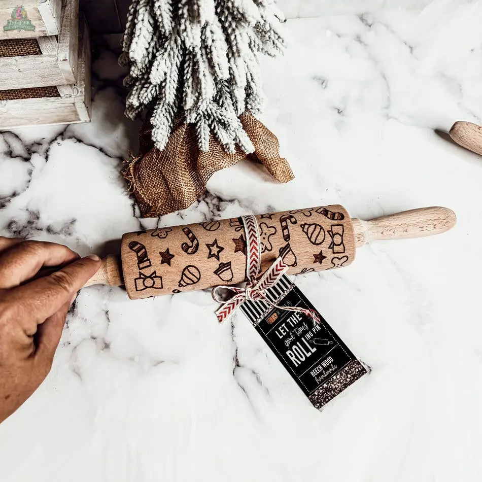 A hand holds a Christmas Rolling Pin Set | 10 Styles, engraved with festive shapes—ideal for holiday baking—beside a small Christmas tree and a tag that says, "Let the good times roll.