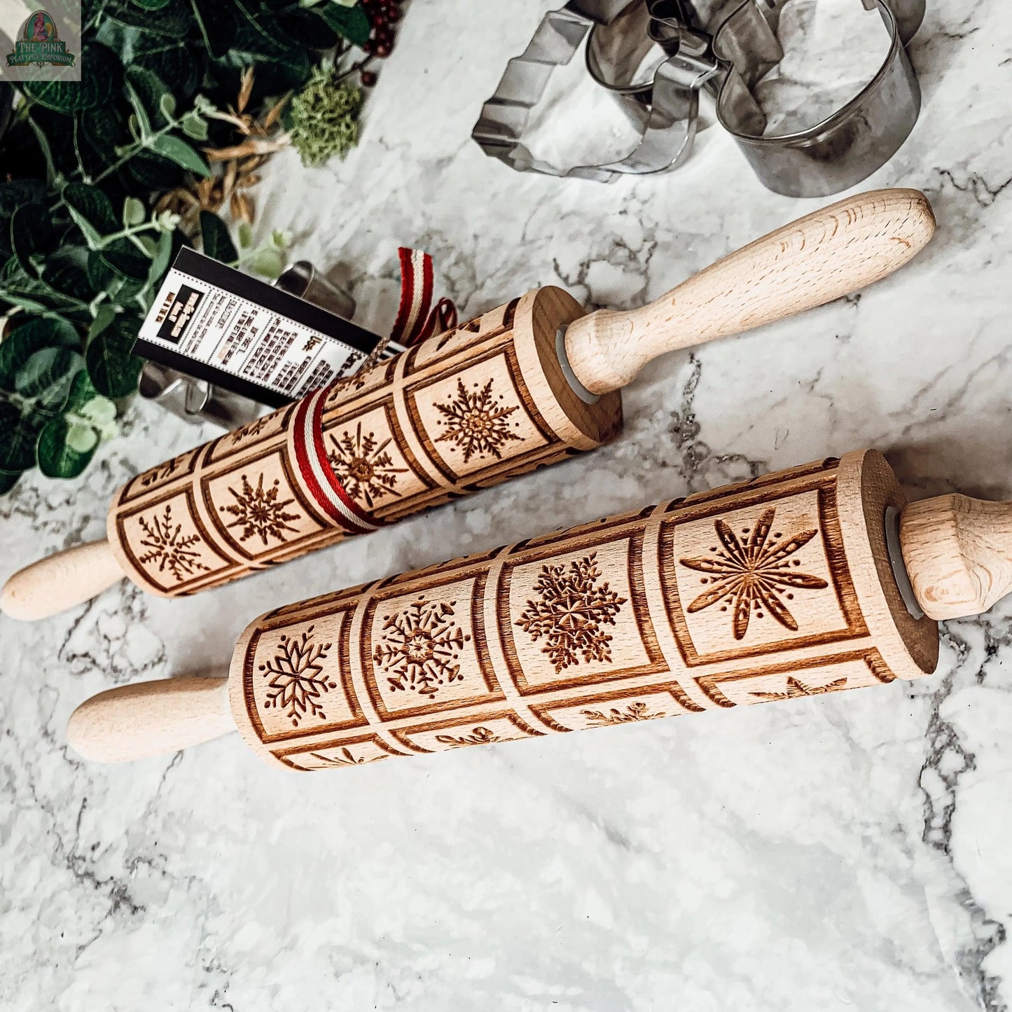 Two Christmas Rolling Pin Set | 10 Styles with engraved snowflake patterns rest on a marble countertop near cookie cutters and green foliage. One, ideal as a baking gift, is wrapped with a red and white ribbon holding a tag.