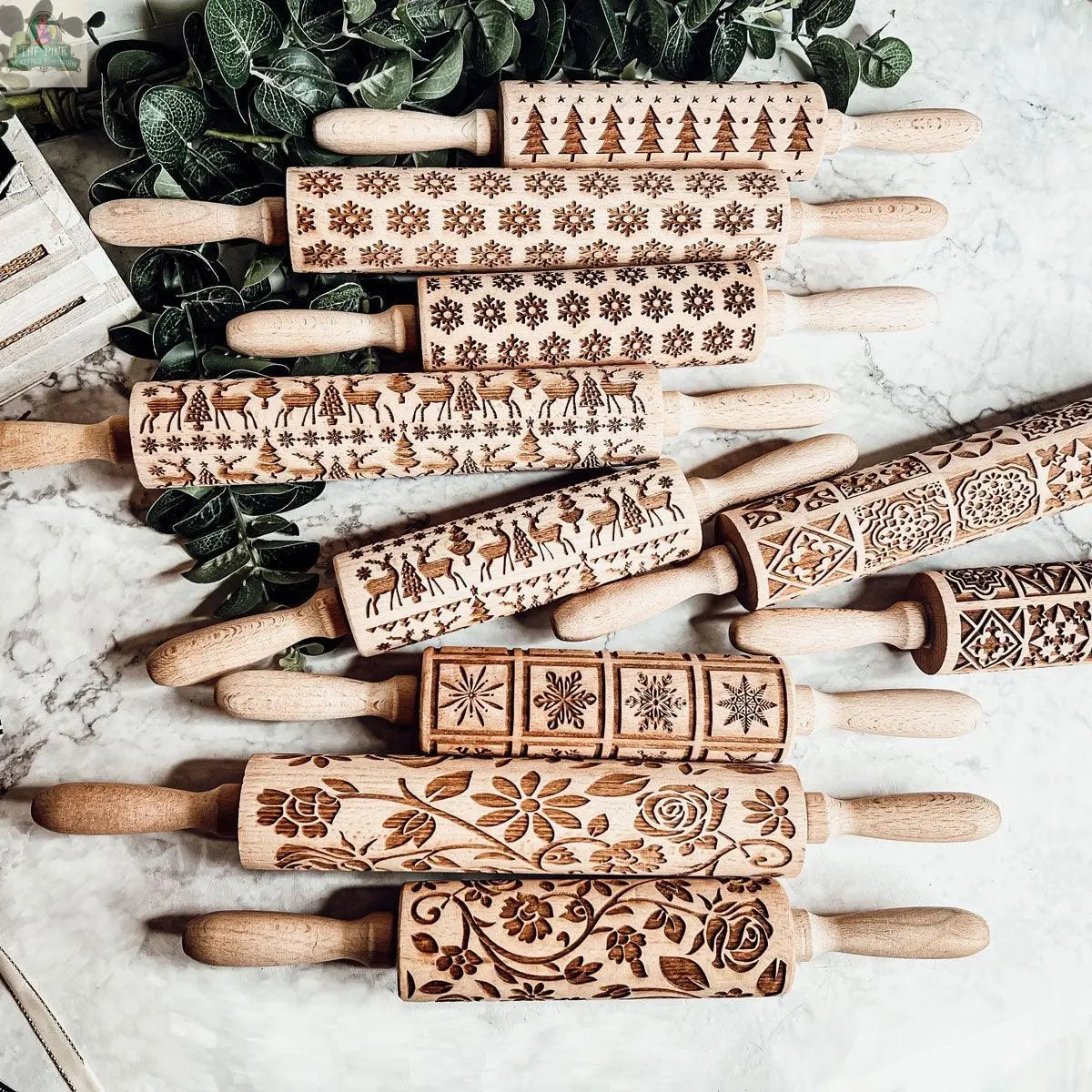 The Christmas Rolling Pin Set | 10 Styles features wooden pins with festive carved designs—trees, snowflakes, reindeer, flowers, and geometric patterns—arranged on marble with greenery, making a delightful gift for baking enthusiasts.