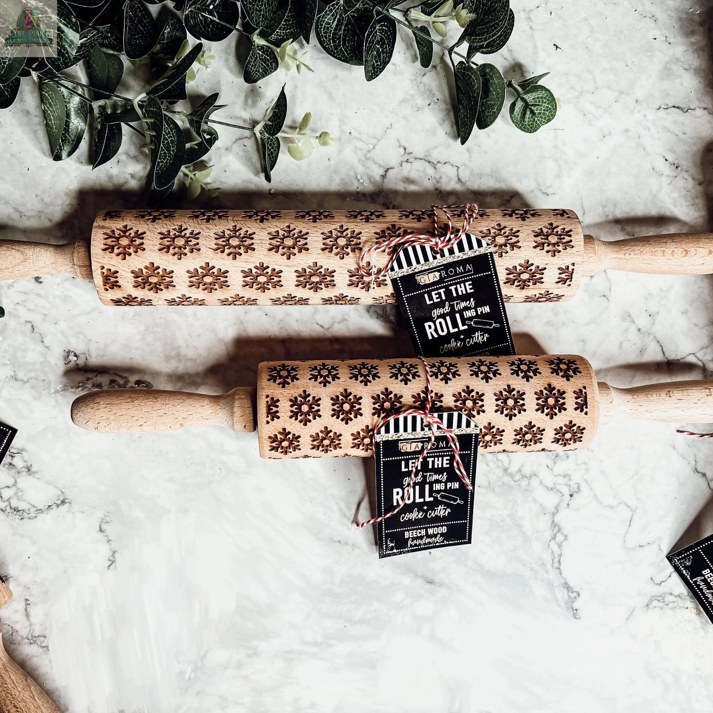 Two Christmas Rolling Pin Set | 10 Styles with carved snowflake patterns rest on marble, each tied with red-and-white twine and a black tag, perfect for holiday baking. Green leafy branches decorate the upper left corner.