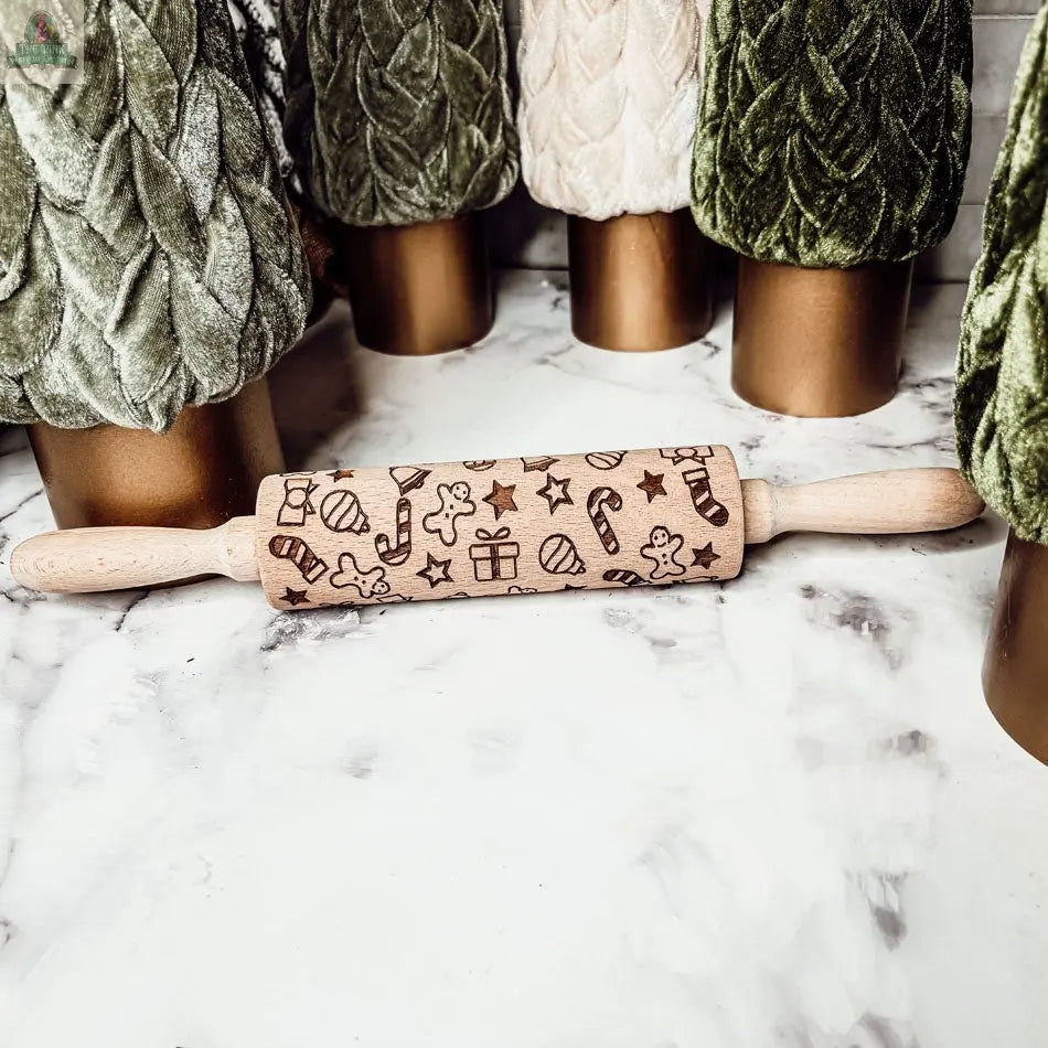 The Christmas Rolling Pin Set | 10 Styles features engraved wooden pins with festive designs, placed on a marble surface amid cookie cutters and velvet-textured decor—perfect for holiday baking fun.