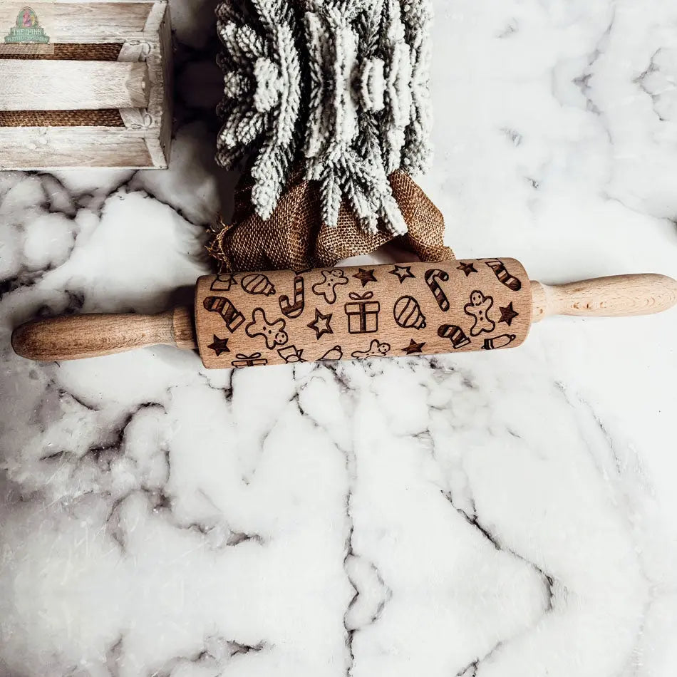The Christmas Rolling Pin Set | 10 Styles features engraved festive designs and rests on a white marble surface with cookie cutters, a frosted leafy bundle, and a wooden crate—making it an ideal, charming baking gift.
