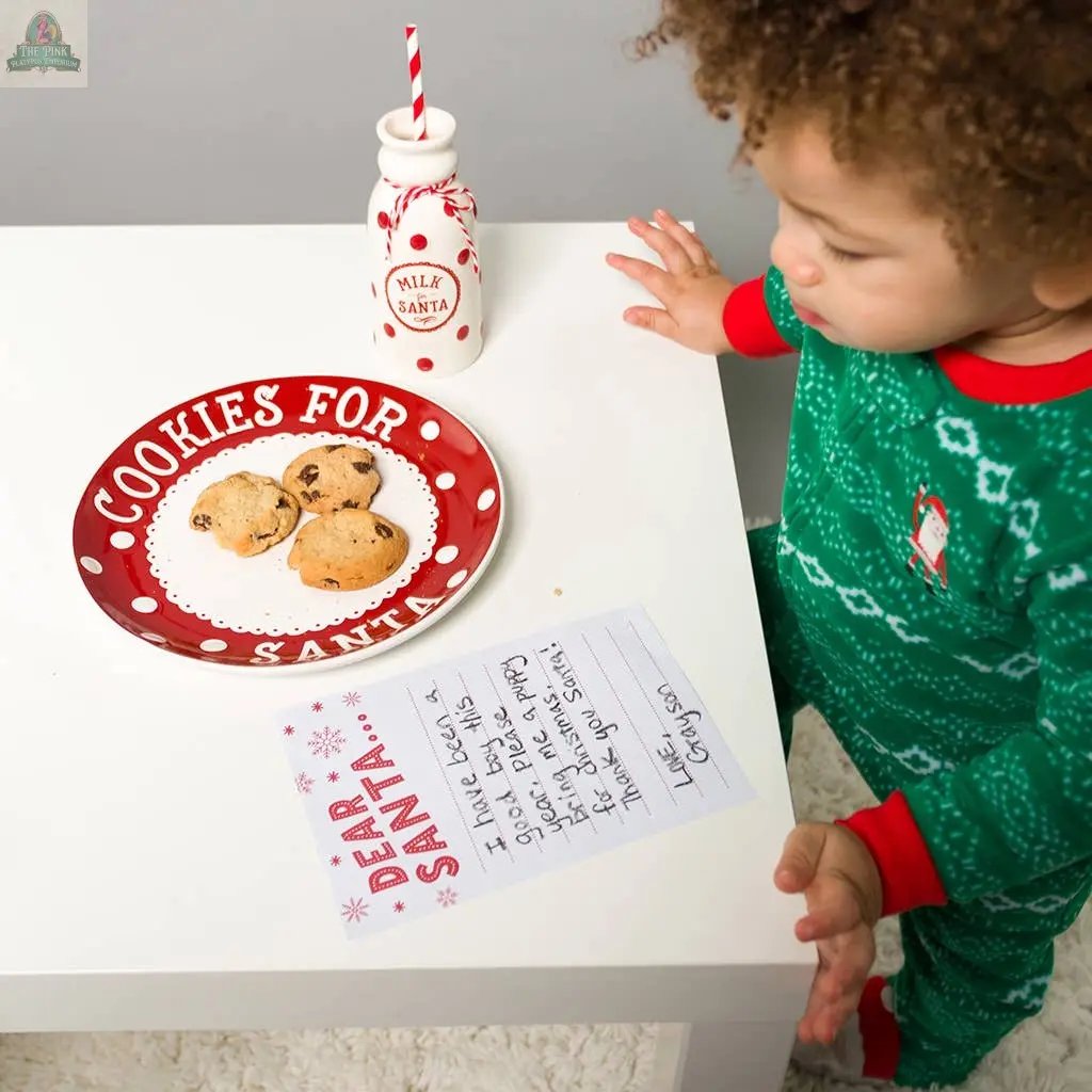 A young child in green Christmas pajamas stands by a white table with the Christmas Santa's Milk Jug, Plate and Dear Santa Notepad Set, featuring a Santa cookie plate, milk jug, and a notepad for writing to Santa.