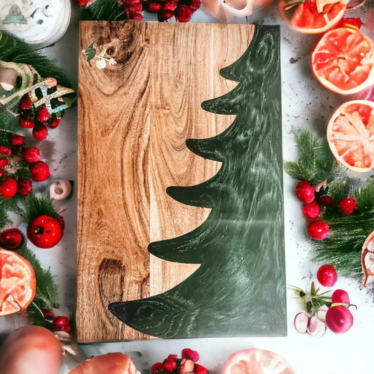 The Christmas Serving Board is a hand-crafted wooden charcuterie board featuring a dark green Christmas tree design on the right, surrounded by festive accents like berries, pine branches, dried citrus slices, and mushrooms on a light background.