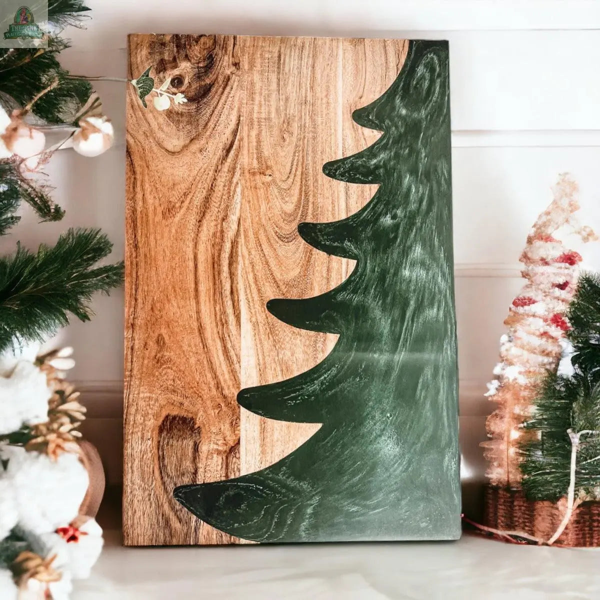 The Christmas Serving Board is a hand-crafted rectangular wooden charcuterie board featuring a green painted Christmas tree silhouette, accented with festive holiday faux greenery and ornaments.