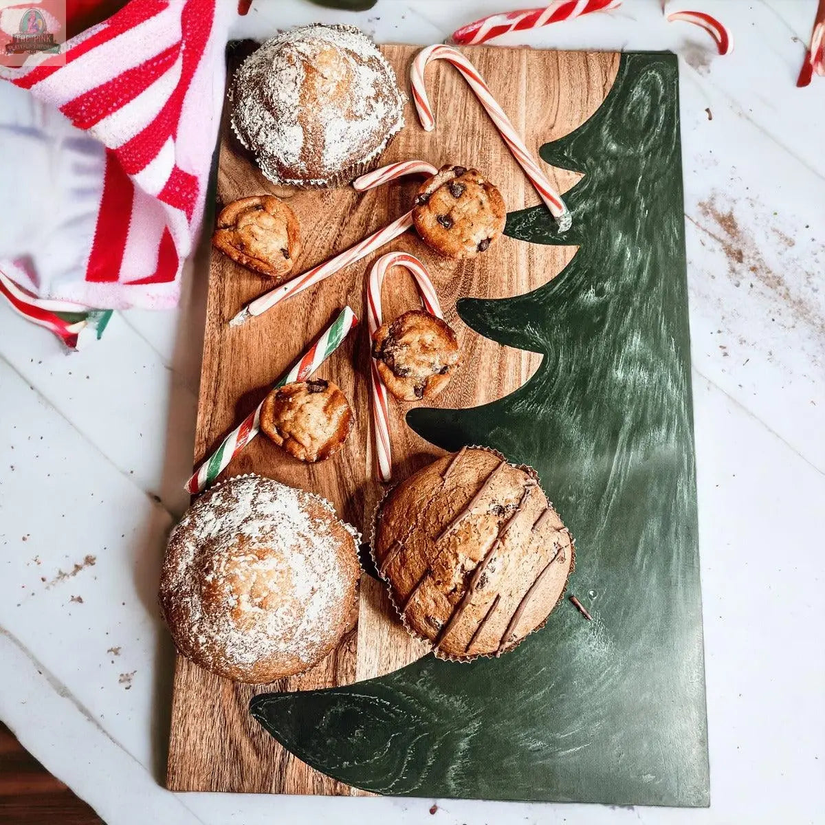 The Christmas Serving Board, featuring a hand-painted green Christmas tree, displays muffins, cookies, and candy canes, accented with a red-and-white striped cloth on the side.