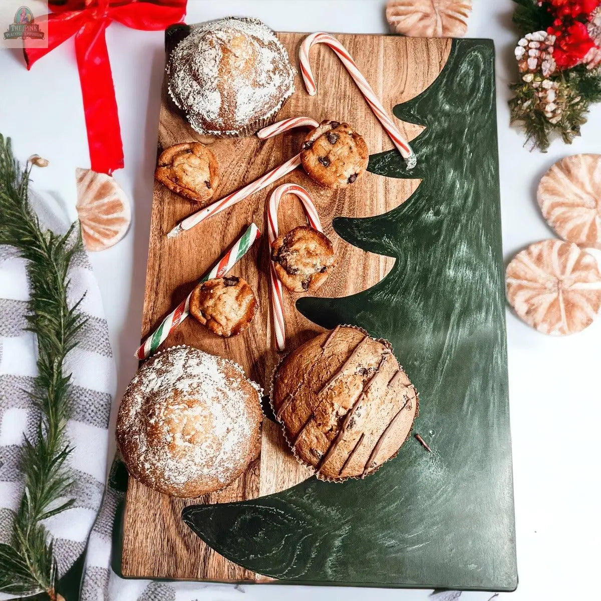 The Christmas Serving Board, featuring a green Christmas tree design, displays muffins, cookies, and candy canes. Festive decorations, a red ribbon, and greenery surround the board, creating a cheerful holiday atmosphere.