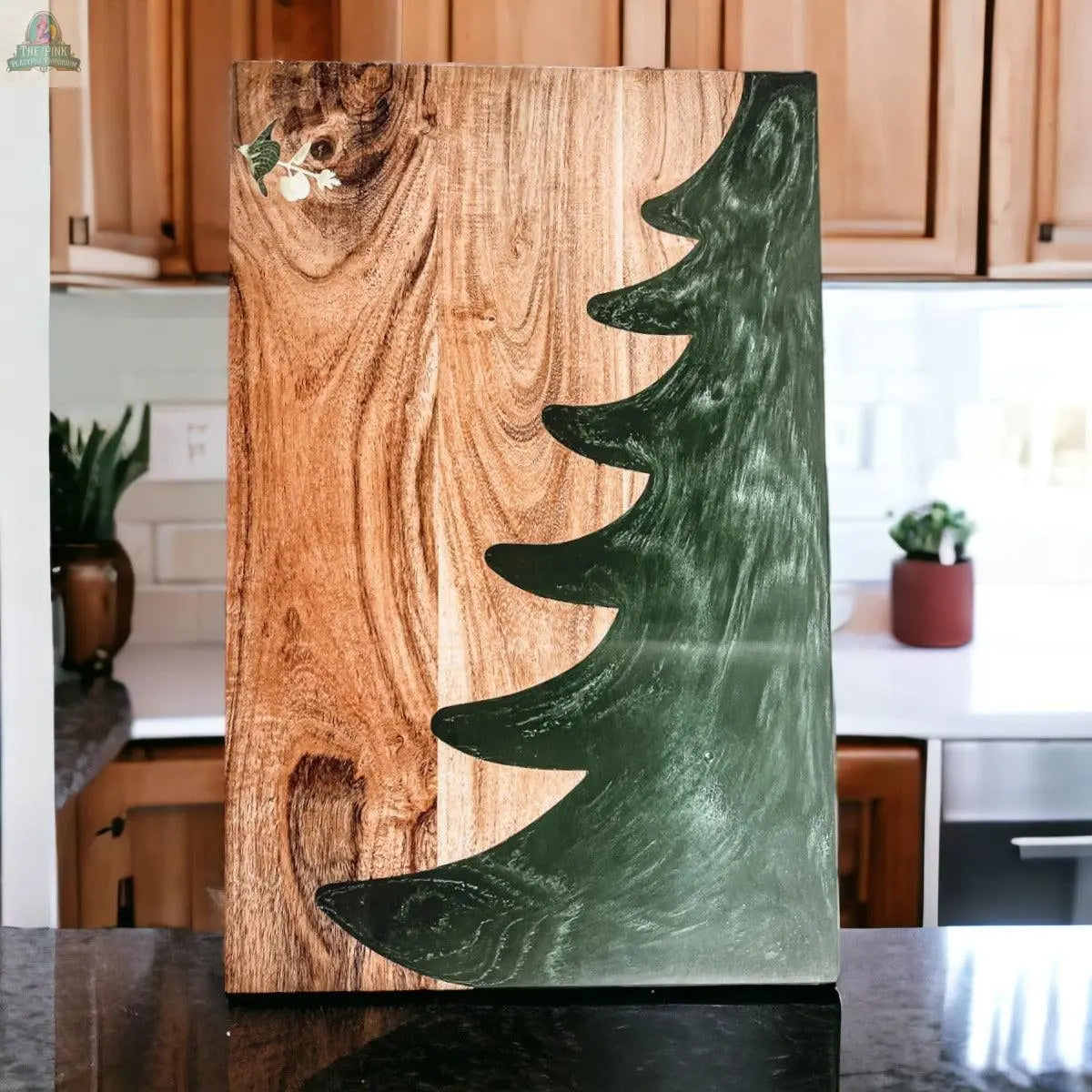 The Christmas Serving Board stands upright on a kitchen counter, featuring a natural wood finish on one side and a green-painted Christmas tree silhouette on the other, with cabinets and plants in the background.