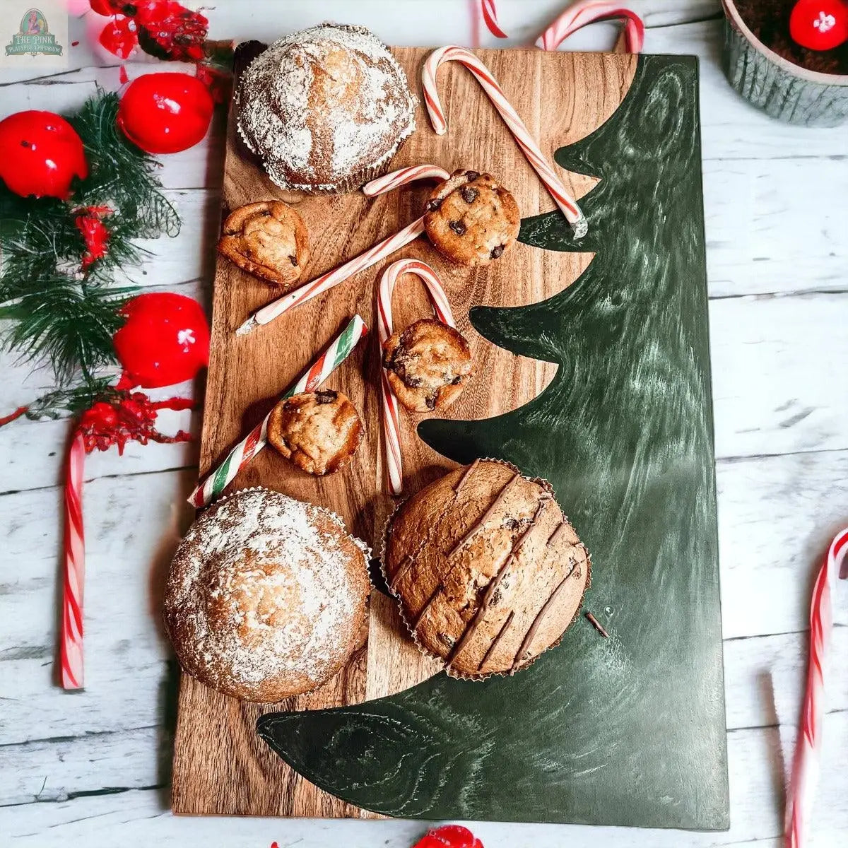 The Christmas Serving Board, shaped like a tree, holds muffins, cookies, and candy canes, while red ornaments and festive evergreen branches with berries decorate the white wooden surface around it.