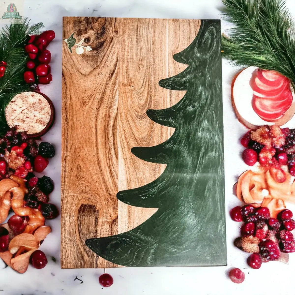 The Christmas Serving Board, featuring a green Christmas tree design, rests on a marble surface surrounded by festive treats, berries, cookies, and pine branches arranged on both sides.