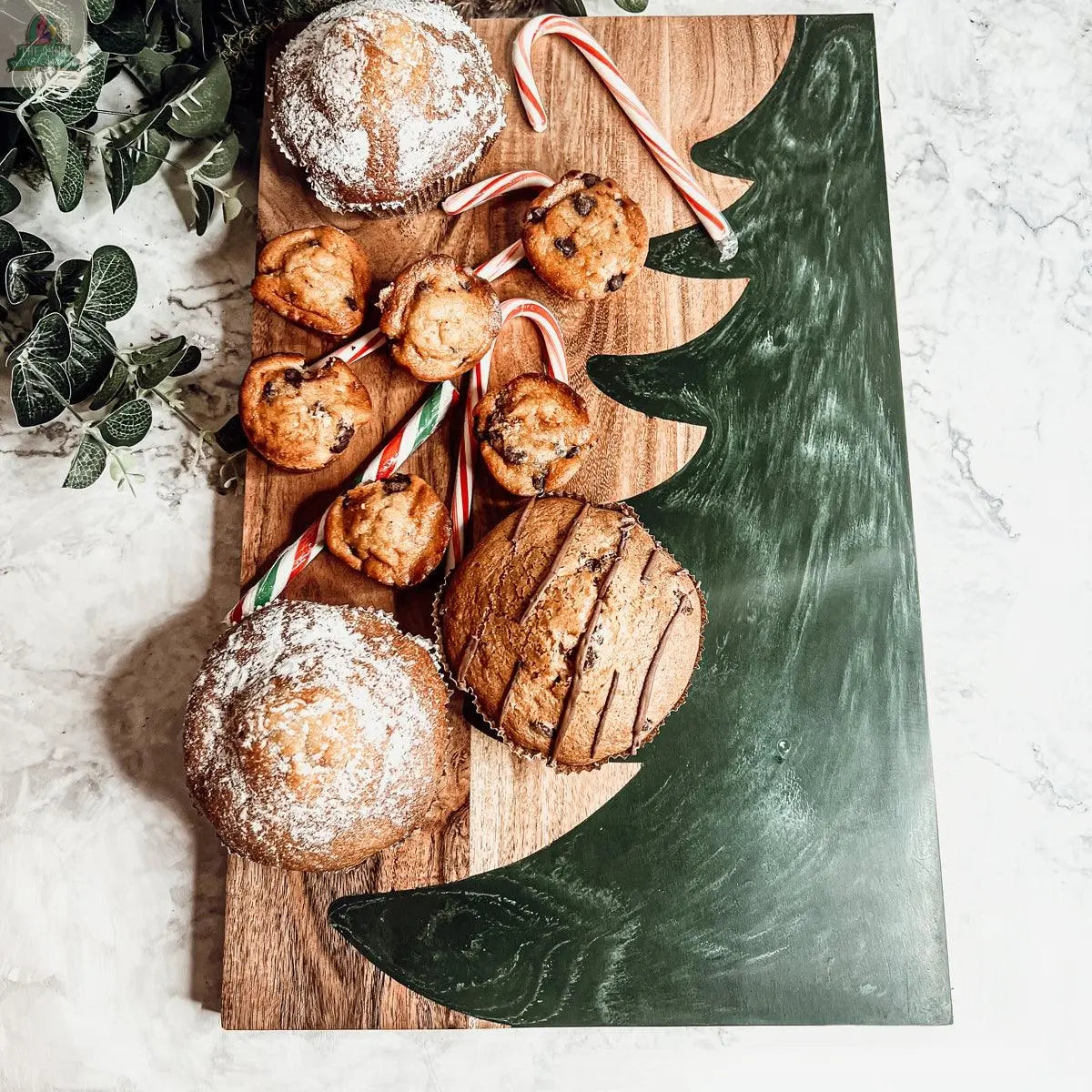 The Christmas Serving Board, shaped and painted like a festive tree, displays muffins, powdered sugar cupcakes, and candy canes on a white marble surface with green leaves in the corner.