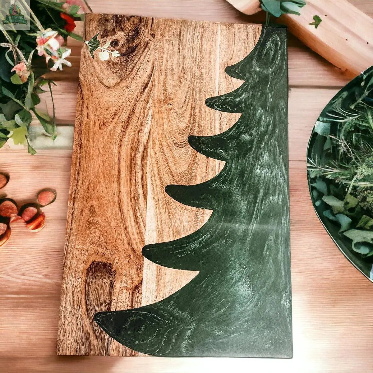 The Christmas Serving Board is a hand-crafted wooden charcuterie board featuring one edge shaped and colored like a green Christmas tree, displayed on a wooden table with leaves, flowers, and chestnuts.
