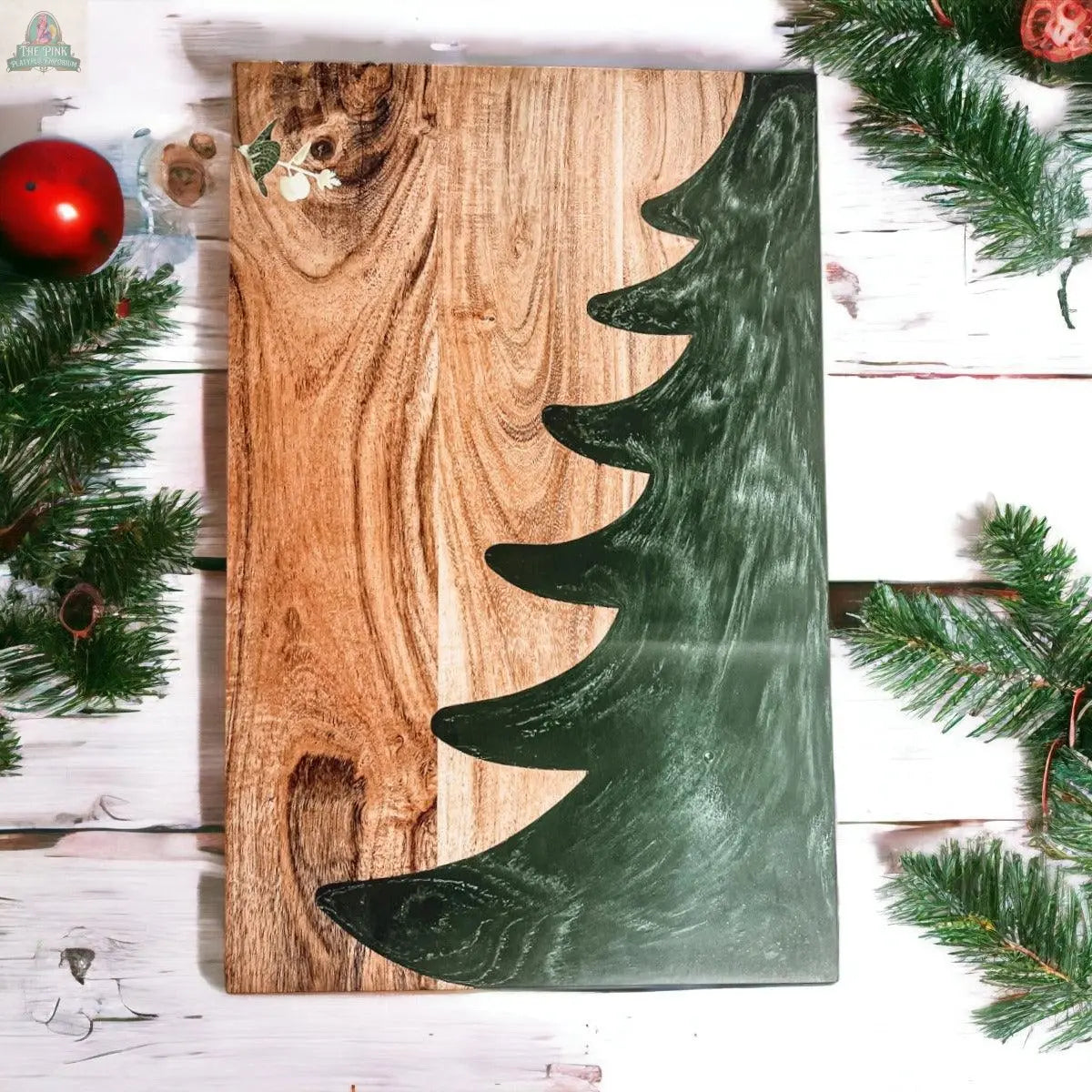 The Christmas Serving Board is a hand-crafted wooden charcuterie board, featuring a painted green Christmas tree silhouette accented with pine branches and red ornaments on a white background.